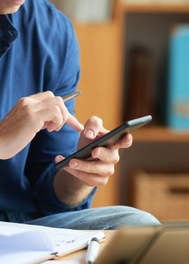 Person in a blue shirt using a smartphone, with documents and a tablet on the desk in a home office setting.